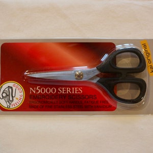 Może przedstawiać: Zbliżenie na parę czarnych nożyczek do haftu z srebrnym ostrzem. Opakowanie jest czerwono-białe i zawiera napis "N5000 Series Embroidery Scissors" oraz "Ergonomically soft handle, fatigue free. Made of fine stainless steel with vanadium." Opakowanie zawiera również napis "Kai Original" i "Made in Japan".