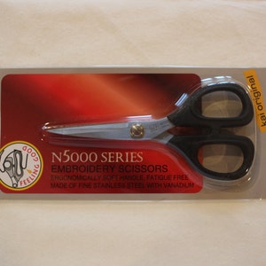 May include: A close-up of a pair of black embroidery scissors with a silver blade. The packaging is red and features the text "N5000 SERIES EMBROIDERY SCISSORS ERGONOMICALLY SOFT HANDLE FATIGUE FREE MADE OF FINE STAINLESS STEEL WITH VANADIUM" and "kai original".