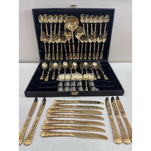 May include: A gold-toned cutlery set arranged in a black storage case with a velvet-lined interior. The set includes various spoons, forks, and knives with ornate handles. Several knives are laid out in front of the case on a flat surface.