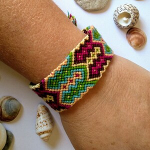 May include: A colourful friendship bracelet with a geometric pattern in shades of pink, green, blue, and brown. The bracelet is woven with a variety of threads and is tied around a person's wrist.