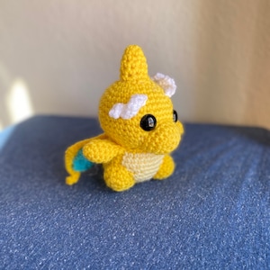 May include: A crocheted yellow and white plush toy with black eyes. The toy is sitting on a blue fabric surface.