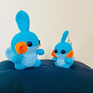 Op de afbeelding: Twee gehaakte blauwe en oranje pluche speelgoedjes. Het grotere speelgoed staat rechtop en het kleinere zit. Beide speelgoedjes hebben zwarte ogen en oranje bekken.