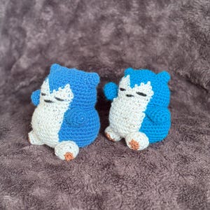 May include: Two handmade crochet figures of a blue and white creature with closed eyes and small arms. The figures are sitting on a brown, textured surface. The creatures have small, tan-colored feet.