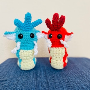 May include: Two crocheted plush toys, one blue and one red, with white accents. They have large black eyes and are standing on a blue surface.