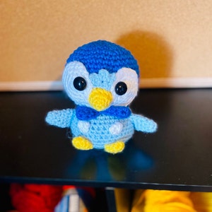 May include: A blue and white crocheted penguin plush toy with yellow beak and black eyes. The penguin is wearing a blue bow tie.