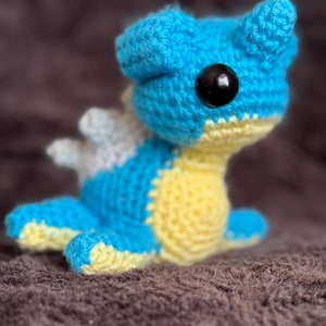Puede incluir: Un peluche de Lapras hecho a mano con ganchillo en azul, amarillo y blanco. El peluche tiene un ojo negro grande y orejas pequeñas. El cuerpo es principalmente azul con detalles amarillos en el vientre y los pies. Las alas son blancas.