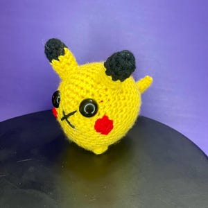 May include: A crocheted yellow Pikachu plush toy with black ears and eyes. The toy has red cheeks and a small tail.