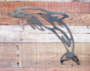 May include: A metal dolphin silhouette wall decor. The dolphin is leaping out of the water with its tail and fins extended. The dolphin is a dark gray color and is mounted on a light brown wood background.