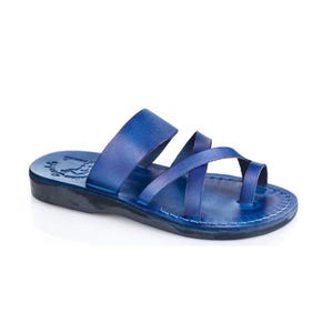 May include: A vibrant blue leather sandal featuring a wide strap across the midfoot, a crisscross strap design over the toes, and a separate toe loop. The flat sole is dark, and the footbed displays a stamped logo near the heel area.