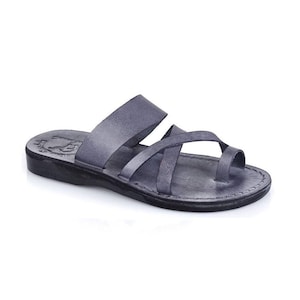 May include: A dark gray leather slide sandal featuring a wide strap across the midfoot and a crisscross strap design that includes a toe loop. The flat sole is black with visible stitching along the edge and a stamped logo on the heel area.
