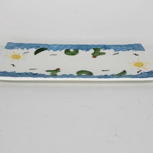 May include: A rectangular white ceramic tray featuring a hand-painted border of blue waves along the top and bottom edges. The center design includes yellow suns, green cacti with small red accents, and black wavy lines scattered across the surface.
