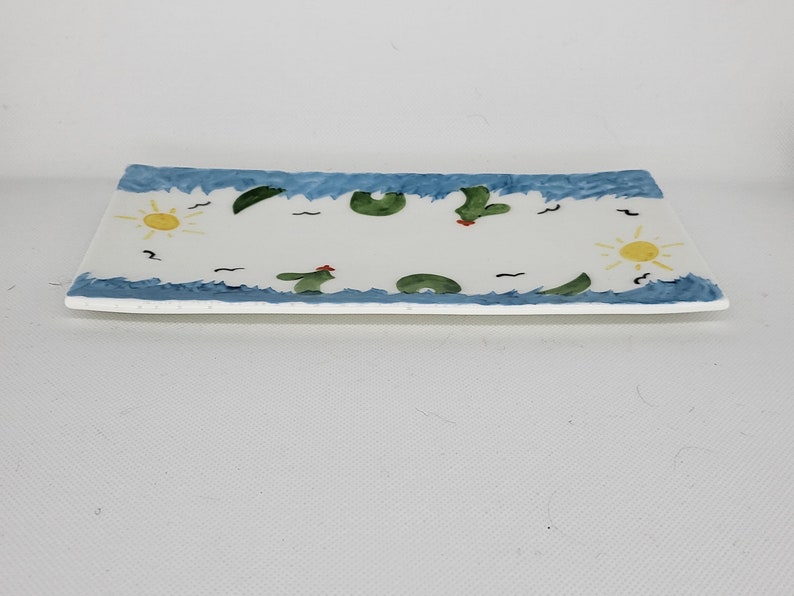 May include: A rectangular white ceramic tray featuring a hand-painted border of blue waves along the top and bottom edges. The center design includes yellow suns, green cacti with small red accents, and black wavy lines scattered across the surface.