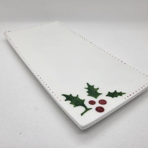 May include: A rectangular white ceramic serving platter with a holly design. The holly features green leaves and red berries. The edge of the platter has a dotted border in a reddish-brown colour.
