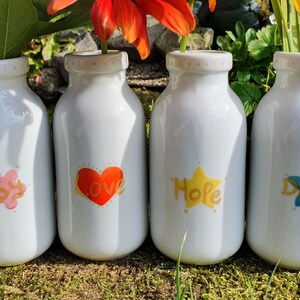 Op de afbeelding: Vier witte keramische vazen met bloemen- en woordontwerpen. Elke vaas heeft een ander ontwerp: een roze wolk met "Joy", een rode hart met "Love", een gele ster met "Hope" en een blauwe bloem met "Dream".