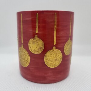 Puede incluir: Una taza cilíndrica de cerámica roja con un acabado texturizado. La taza está adornada con diseños de adornos navideños dorados y estilizados. Los adornos están suspendidos de líneas doradas, creando un tema festivo navideño.
