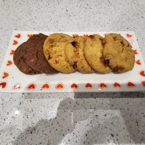 May include: A rectangular white serving platter with a red heart pattern holds an assortment of cookies. There are chocolate and golden brown cookies. The cookies are arranged on the platter, ready to be served.