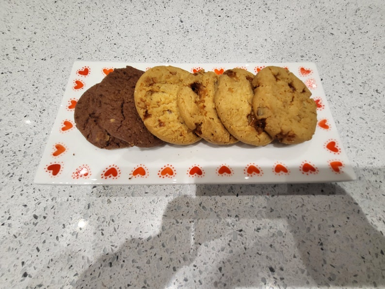 May include: A rectangular white serving platter with a red heart pattern holds an assortment of cookies. There are chocolate and golden brown cookies. The cookies are arranged on the platter, ready to be served.