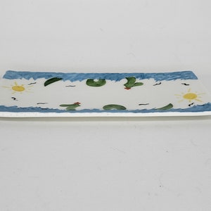May include: A rectangular ceramic serving tray with a white center and a blue wavy border. The design features hand-painted yellow suns, green cacti with small red accents, and black bird silhouettes scattered across the surface of the tray.