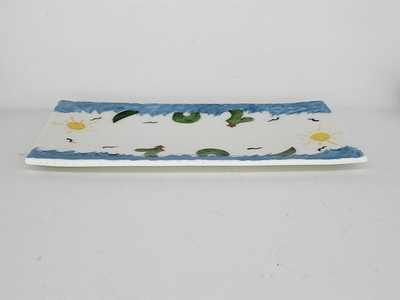 May include: A rectangular ceramic serving tray with a white center and a blue wavy border. The design features hand-painted yellow suns, green cacti with small red accents, and black bird silhouettes scattered across the surface of the tray.