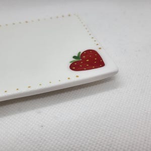 May include: A white rectangular ceramic plate featuring a red strawberry design. The strawberry is heart-shaped with green leaves and yellow seeds. The plate's edge is decorated with gold dots.