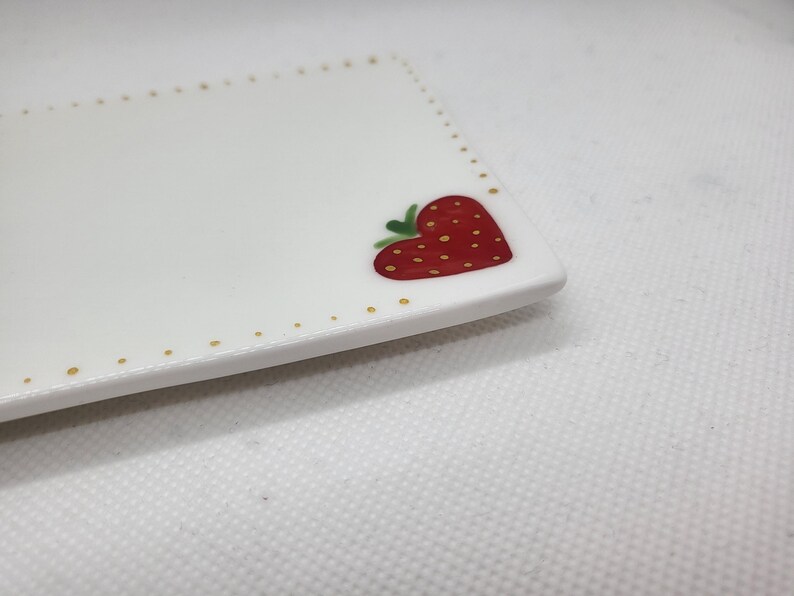 May include: A white rectangular ceramic plate featuring a red strawberry design. The strawberry is heart-shaped with green leaves and yellow seeds. The plate's edge is decorated with gold dots.