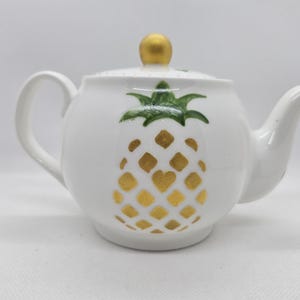 May include: A white ceramic teapot featuring a painted pineapple design on the front. The pineapple has a green leafy top and a body composed of gold diamond-shaped patterns. The teapot includes a matching lid with a round gold knob on top.