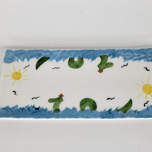 May include: A rectangular white ceramic tray featuring a hand-painted border of blue waves along the top and bottom edges. The design includes green cactus shapes, yellow suns on the left and right sides, and small black bird silhouettes.