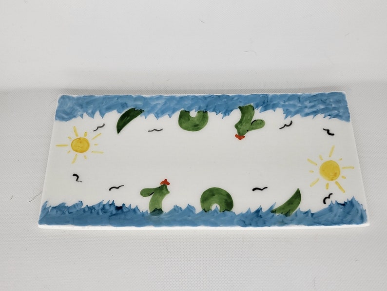 May include: A rectangular white ceramic tray featuring a hand-painted border of blue waves along the top and bottom edges. The design includes green cactus shapes, yellow suns on the left and right sides, and small black bird silhouettes.