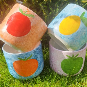 May include: Four hand-painted ceramic planters with fruit designs. The planters are stacked, showcasing a tomato, lemon, and green tomato. The planters have a textured finish and are set on a bed of green grass.