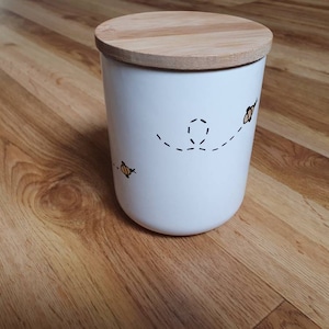 May include: A white ceramic canister with a wooden lid. The canister has a hand-drawn design of bees with dotted flight paths. The lid is made of light-coloured wood. The canister is cylindrical in shape.
