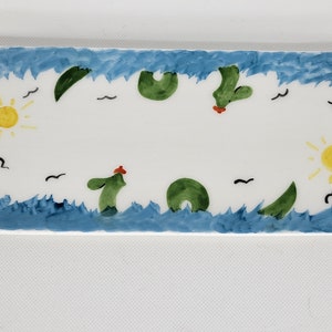 May include: A rectangular white ceramic tray featuring a hand-painted scene with blue wavy borders representing water. The design includes two yellow suns, green cactus-like shapes, and small black bird silhouettes scattered across the surface.