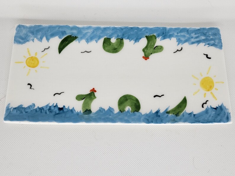 May include: A rectangular white ceramic tray featuring a hand-painted scene with blue wavy borders representing water. The design includes two yellow suns, green cactus-like shapes, and small black bird silhouettes scattered across the surface.