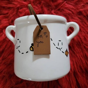 May include: White ceramic mug with two handles, decorated with a bee and dotted flight path design. A brown tag with the handwritten text "For you" and two hearts hangs from the handle. The mug sits on a red, textured surface.