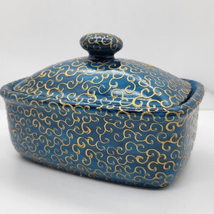 Può includere: Un contenitore in ceramica blu con coperchio, ornato da un motivo a spirali dorate. Il contenitore ha una forma rettangolare con bordi arrotondati e un piccolo pomello sulla parte superiore del coperchio. Il design è uniforme.