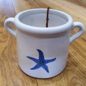 May include: White ceramic pot with two handles and a blue starfish design. The pot has a brown leather strap inside. The pot is decorated with small white dots around the rim. This decorative pot is perfect for plants.