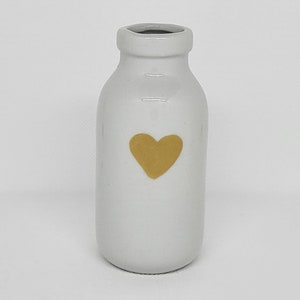 Op de afbeelding: Een kleine, witte keramische vaas met een gouden hartontwerp. De vaas heeft de vorm van een melkfles met een smalle hals en een iets bredere basis. Het hart is in het midden aan de voorkant geplaatst.