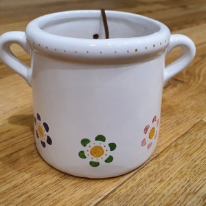May include: White ceramic pot with two handles and a floral design. The pot features colourful flowers in purple, green, and pink. The rim is decorated with small gold dots. A brown string is visible inside the pot.