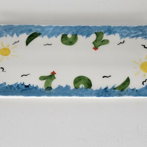 May include: A rectangular ceramic tray featuring a hand-painted scene with blue waves along the top and bottom edges. The white center displays green sea creatures, yellow suns on the left and right sides, and small black birds scattered throughout.