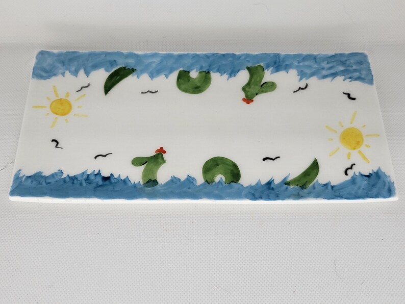 May include: A rectangular ceramic tray featuring a hand-painted scene with blue waves along the top and bottom edges. The white center displays green sea creatures, yellow suns on the left and right sides, and small black birds scattered throughout.