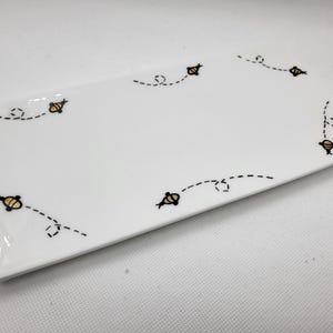 May include: A rectangular white ceramic plate with a glossy finish. The plate is decorated with a repeating pattern of small yellow and black bees with dotted flight paths. The bees are evenly spaced around the edges of the plate.