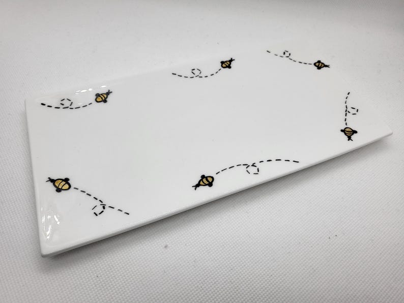 May include: A rectangular white ceramic plate with a glossy finish. The plate is decorated with a repeating pattern of small yellow and black bees with dotted flight paths. The bees are evenly spaced around the edges of the plate.