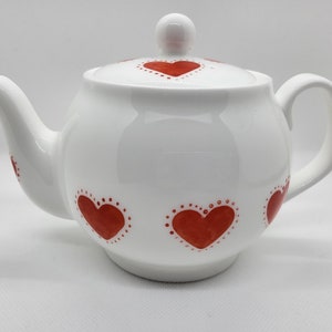 May include: White ceramic teapot with a lid and handle. The teapot is decorated with red hearts outlined with small white dots. The spout and lid handle are also white.