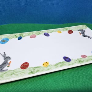 Peut inclure: Un plat rectangulaire en céramique blanche sur le thème de Pâques. Le plat présente une bordure d'herbe verte et d'œufs de Pâques colorés. Un lapin gris tenant une carotte est sur le côté gauche. Un oiseau gris est sur le côté droit.