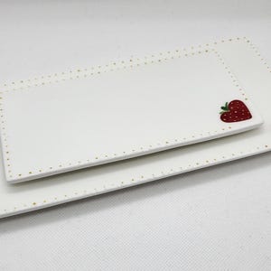 May include: Two rectangular white ceramic serving platters. The edges are dotted with gold. Each platter has a red strawberry heart design in the corner. The platters are nested together.