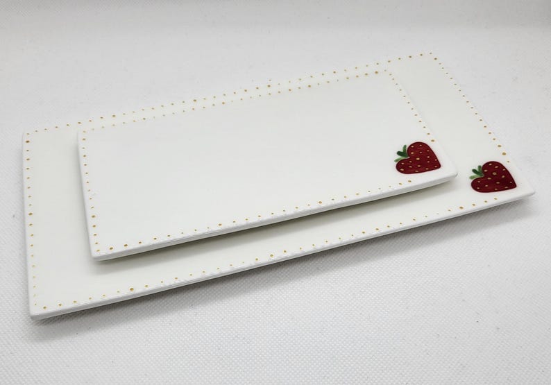 May include: Two rectangular white ceramic serving platters. The edges are dotted with gold. Each platter has a red strawberry heart design in the corner. The platters are nested together.
