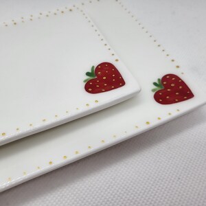 May include: Two white rectangular ceramic plates with a strawberry heart design. The strawberries are red with yellow seeds and green leaves. The plates have a border of small gold dots.