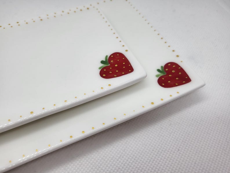 May include: Two white rectangular ceramic plates with a strawberry heart design. The strawberries are red with yellow seeds and green leaves. The plates have a border of small gold dots.