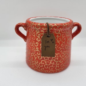 May include: A red ceramic pot with gold swirl patterns and two handles. A brown tag with the words "For you" and a heart is attached. The interior of the pot is white.