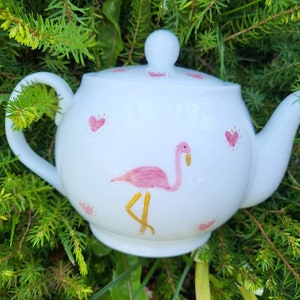 May include: White ceramic teapot decorated with a flamingo motif. The teapot has a pink flamingo with gold legs and pink hearts. It features a curved handle, a spout, and a lid with a small knob. The teapot is set against a backdrop of green foliage.