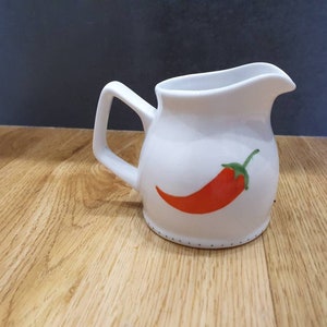 May include: White ceramic creamer with a red chilli pepper design. The creamer has a curved handle and a spout for easy pouring. The chilli pepper is orange with a green stem. The creamer is on a wooden surface.